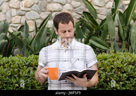 Mann in einem weißen Plaid Shirt Lesen der Bibel außerhalb halten sie eine orange Kaffeebecher mit Pflanzen im Hintergrund. Stockfoto