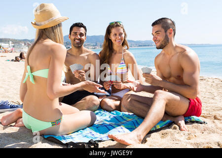 Vier lächelnden Personen Karten sitzen auf Sand Sommer Strand Stockfoto