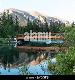 Eine hölzerne Fußgängerbrücke über den Bow River mit Pinienwald und Rocky Mountains mit Reflexionen an Canmore Banff National Park, Alberta, Kanada Stockfoto