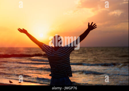 Die Silhouette eines Mannes, der seine Hände zum Himmel bei Sonnenuntergang am Strand und das Beten zu Gott Stockfoto