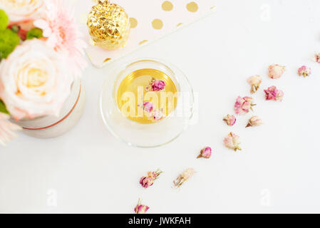 Eine Tasse gesunden Kräutertee mit getrockneten Rosen. Schöne frische Blumen, goldene Ananas, Notebook auf hellem Marmor Tabelle, Ansicht von oben. Rosa Rosen und gerb Stockfoto