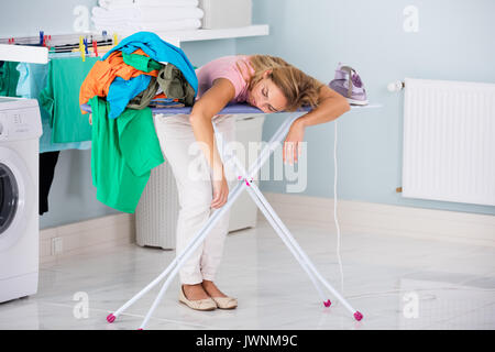 Junge müde Frau Schlafen auf dem Bügelbrett Neben der Kleidung zu Hause Stapel Stockfoto