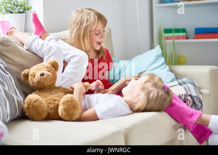 Hübsche blonde Mädchen lesen Abenteuer Geschichte laut, während ihre kleine Schwester lag auf dem Sofa und hören sie mit Interesse auf, Interieur des gemütlichen Livi Stockfoto