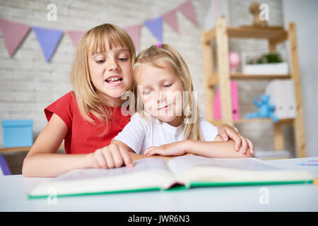 Portrait von der hübschen blonden Mädchen sie sanft Umarmen kleine Schwester während Märchen vorlesen, Interieur der schönen Schlafzimmer auf Hintergrund Stockfoto