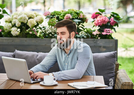 Porträt des zeitgenössischen Bärtigen mit Laptop arbeiten bei Tisch Mann in gemütlicher Café Lounge im Freien Stockfoto