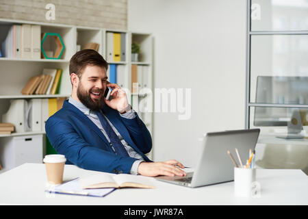 Portrait von erfolgreichen bärtigen Geschäftsmann sprechen per Telefon und lächelnd während der Arbeit am Schreibtisch im Büro Stockfoto