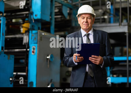 Portrait von älteren Werk Eigentümer tragen, hardhat auf Tour in der modernen Fabrik in die Kamera schauen und lächeln, die Zwischenablage Stockfoto