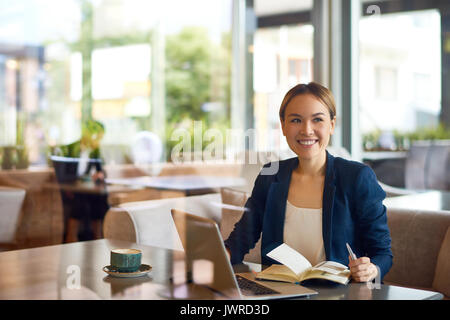 Taille-up Portrait von hübschen Asiatischen Geschäftsfrau mit charmanten Lächeln sitzt im gemütlichen kleinen Café und arbeiten an vielversprechenden Projekt, die Ansicht durch panorami Stockfoto