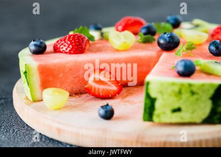 Schnitt in Wassermelone Pizza mit Obst: Erdbeeren, Kiwis, Weintrauben, Heidelbeeren und Minze, auf Holzbrett. Dunkler Hintergrund, Nahaufnahme Stockfoto