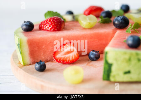 Schnitt in Wassermelone Pizza mit Obst: Erdbeeren, Kiwis, Weintrauben, Heidelbeeren und Minze, auf Holzbrett. Weißer Hintergrund Stockfoto