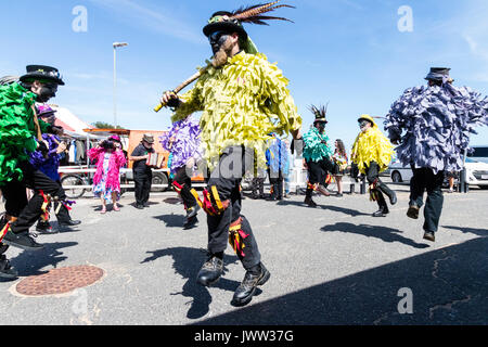 Traditionelle englische Volkstänzer, Motley Morris Team, tanzen. Holding Holzpfähle, die Sie tragen, tatter Jacken in verschiedenen Farben und haben Gesichter geschwärzt. Strahlender Sonnenschein. Stockfoto