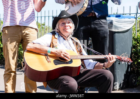 Ältere Frau sitzt auf der Stuhl auf der Strandpromenade in Broadstairs während des Folk Woche Festival. Strumming Akustik Gitarre, trägt Cowboys hat und nach vorne. Zeigt. Stockfoto