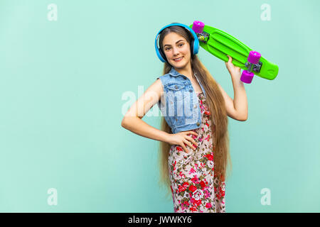 Die happy schöne Langhaarige blonde Mädchen im blauen Kleid und Kopfhörer, Spaß mit grünen Kunststoff penny Skateboard in gekreuzten Händen onder Kopf, Stockfoto