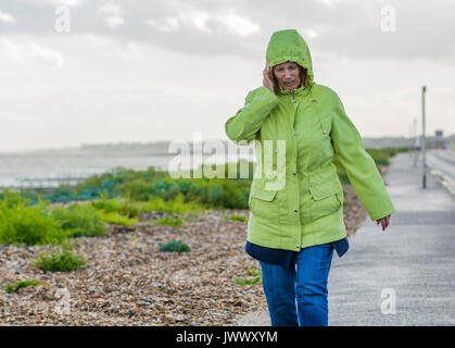 Ältere Frau zu Fuß entlang der Strandpromenade in bewölkt nassen und windigen Wetter, halten Ihre Ohren warm. Stockfoto