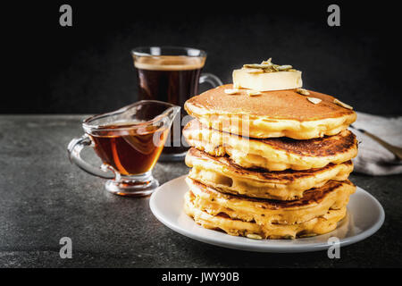 Herbstliche traditionelles Essen. Stapel von Kürbis Pfannkuchen mit Butter, Kürbiskerne und Ahornsirup. Mit einer Tasse Kaffee. Auf einem schwarzen Steintisch. Kopieren Sie sp Stockfoto