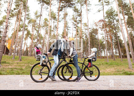 Schöne junge Familie mit zwei Töchtern im Fahrrad sitze in warme Kleidung Radfahren außerhalb im Herbst Natur, Vater Mutter küssen. Stockfoto