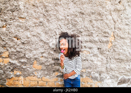 Schöne afrikanische amerikanische Mädchen mit lockigem Haar draußen tragen Gestreifte t-shirt lollipop Essen, gegen alte Betonwand stehend. Stockfoto