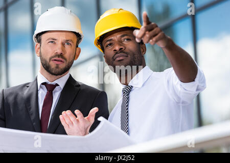 Professionelle Architekten in hardhats Arbeiten mit Blueprint außerhalb modernes Gebäude Stockfoto