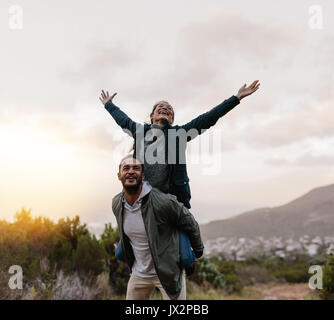 Portrait des jungen Mannes huckepack seine Freundin. Mann, der Frau mit ihren Armen angehoben. Paar selbst in der Landschaft zu genießen.