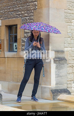 Frau, die an einem regnerischen Tag in Oxford, Oxfordshire, Großbritannien, im August ein Mobiltelefon unter einem Regenschirm benutzt Stockfoto