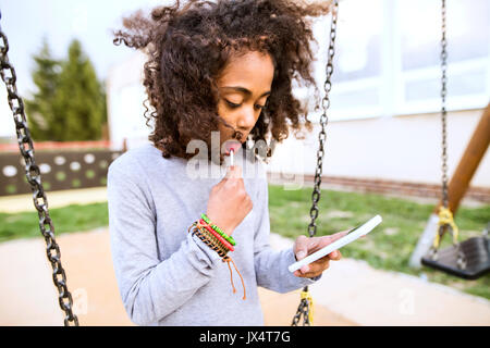 Schöne afrikanische amerikanische Mädchen auf Schaukel mit smart phone schreiben Sms oder etwas zu lesen, Lollipop essen. Stockfoto