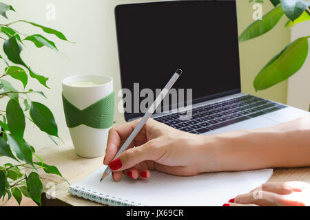 Eine Frau mit hellen Maniküre schreibt in Bleistift im Tagebuch. Neben der Tabelle wird ein Laptop, eine Tasse Wasser mit Zitrone und grünen Pflanzen. Das Konzept o Stockfoto