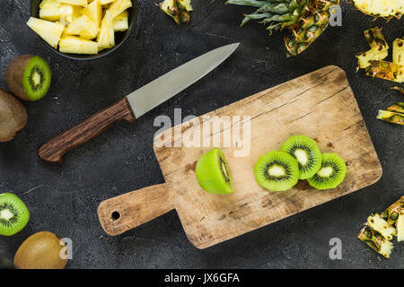 Schneiden frische Kiwi und Ananas auf Holzbrett auf dunklem Hintergrund. Ansicht von oben. Kochen Konzept Stockfoto