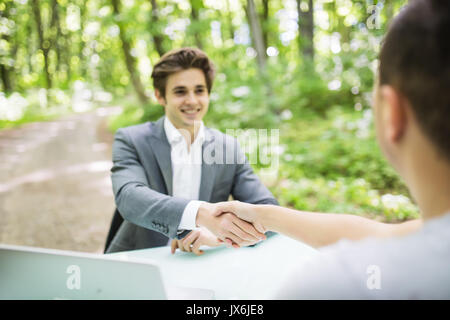 Manager im Anzug begrüßt mit Handshake an Interview neuer Mann in seinem Büro Tisch im Green Park Wald. Geschäftskonzept. Stockfoto