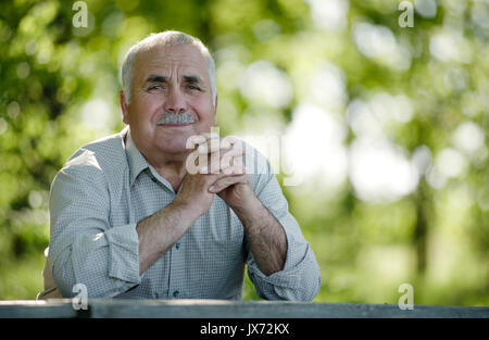 Freundlich lächelnd älterer Mann sitzt entspannt zu einem rustikalen Holztisch im Garten in die Kamera schaut mit einem Lächeln Stockfoto