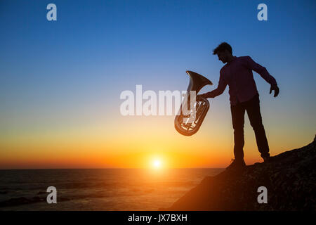 Silhouette des Musikers mit der Tuba auf felsigen Küste bei Sonnenuntergang. Stockfoto