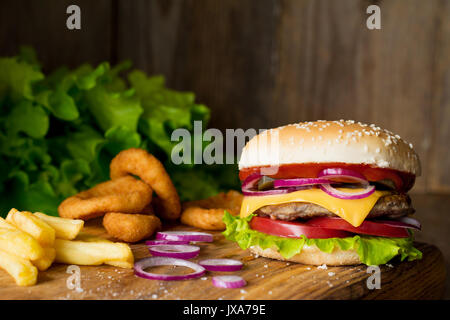 Cheeseburger, Pommes frites und Zwiebelringe auf Holz Schneidebrett über Holz- Hintergrund. Detailansicht, selektive konzentrieren. Fast food Konzept Stockfoto