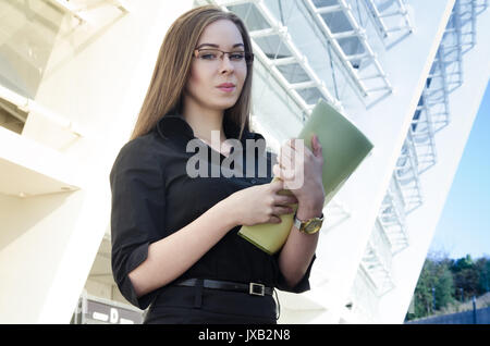Attraktive Geschäftsfrau in Schwarz und Tragen einer Brille mit einem gelben Ordner mit Papieren gekleidet. Listig lächelnd in die Kamera schaut Stockfoto