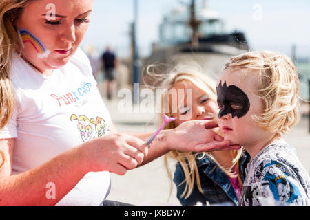 Junge kaukasier Kind, Mädchen, 5-6 Jahre alt, saß in der Sonne in Ihr Gesicht mit einer Art Batman Maske, während ihre Schwester, 6-7 Jahre, Uhren gemalt. Stockfoto