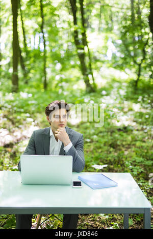 Junge schöne business Mann am Laptop im Büro Tabelle arbeiten mit Hand am Kinn Denken in Green Park. Geschäftskonzept. Stockfoto