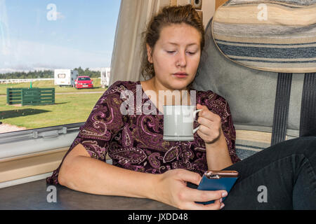 Hübsche junge Frau trinkt Tee oder Kaffee aus einem Becher in einem Wohnmobil auf einem Campingplatz. Exeter, Devon, England, UK. Stockfoto