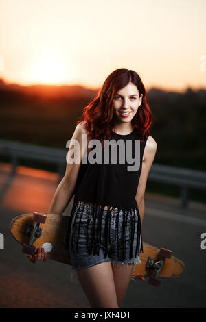 Urban modische Mädchen mit Longboard posiert im Freien auf dem Weg zum Sonnenuntergang. Stockfoto