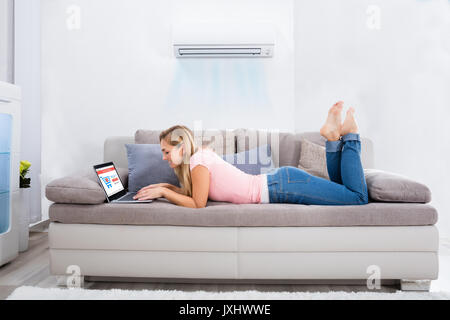 Junge Frau liegen unter der Klimaanlage auf die Couch mit Laptop zu Hause Stockfoto