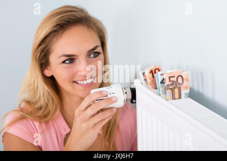 Junge lächelnde Frau Einstellung Thermostat am Kühler mit dem gleichen Geld für Heizkosten Stockfoto