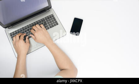 Hände auf einem Laptop mit einer auf einem weißen Tisch mobile beiseite gelegt, gehalten. Ein mockup einer arbeitenden Person auf einem Laptop. Stockfoto