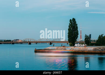 Riga, Lettland. Die konkrete Böschung am Fluss Daugava mit Namen Ortsschild und ruhenden Menschen auf. Radio TV Tower, Stein und Eisenbahnbrücken Backgr Stockfoto