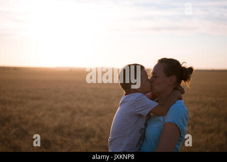 Happy Mother Holding Baby auf ein Weizenfeld im Sonnenlicht, Familie in einem Weizenfeld auf den Sonnenuntergang, junge schöne Frau Küsse auf die Stirn von ihr beleuchteten Stockfoto