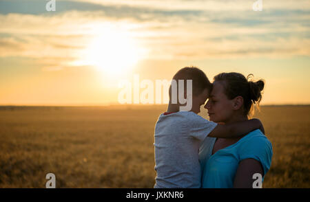 Happy Mother Holding Baby auf ein Weizenfeld im Sonnenlicht, Familie in einem Weizenfeld auf den Sonnenuntergang, junge schöne Frau Küsse auf die Stirn von ihr beleuchteten Stockfoto