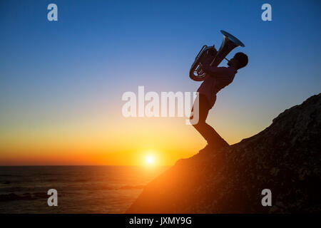 Silhouette des Musikers mit Tuba auf felsigen Küste bei Sonnenuntergang. Stockfoto