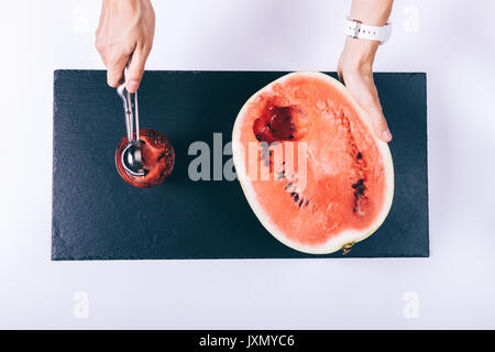 Frauen Hand in Scheiben geschnittenen Wassermelone mit einem Löffel und legen Sie die Stücke in einem Glas auf einem weißen Tisch Ansicht von oben Stockfoto