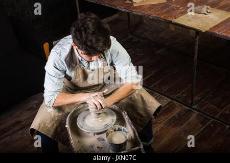 Keramik, Werkstatt, Keramik art concept-Master bildet die Lehm auf Potter's Rad mit Händen, Mann beugte sich über die Arbeit, einen männlichen formt ein utensil in der Nähe von hölzernen Tisch, Mann in Schürze und Shirt, Ansicht von oben. Stockfoto