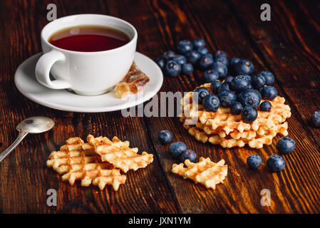 Tasse Tee mit Blaubeeren auf der Oberseite des Belgischen Waffeln Stack und andere verstreut auf Holz- Hintergrund. Stockfoto