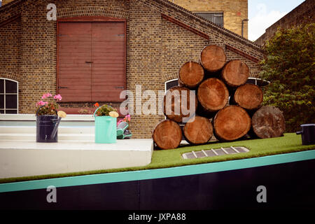 Stapel von Rundholz und Gießkannen mit Blumen auf einem schmalen Boot auf einer London Canal. Querformat. Stockfoto