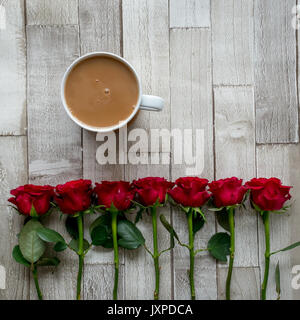 Tasse Tee mit Milch und einer Reihe von roten Rosen auf einem Holzbrett. Flach. Quadratisches Format. Stockfoto