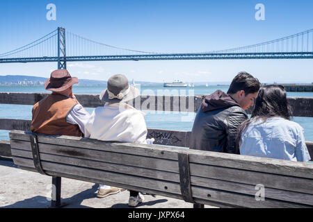 Paare sitzen auf einer Bank mit Blick auf die Oakland Bay Bridge im Hintergrund. Ein junges Paar, ein älterer. Lage: Ferry Building, San Francisco Stockfoto