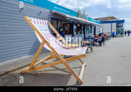 Largs, Schottland, Großbritannien. 17 August, 2017. UK Wetter: Menschen außerhalb der Fisch arbeitet, ein Seafood Restaurant an der Promenade zu bestellen, eine riesige Liegestuhl im Vordergrund sitzen, an einem Tag des sonnigen Abschnitten und gelegentlichen schweren Duschen am Meer an der Westküste von Schottland. Credit: Skully/Alamy leben Nachrichten Stockfoto